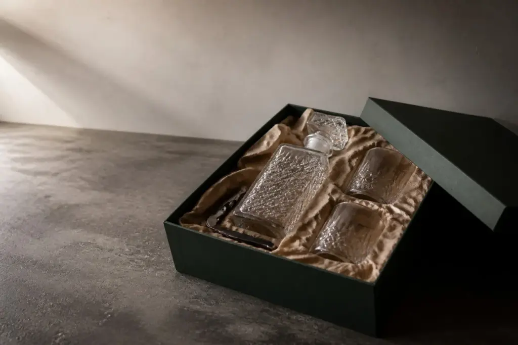 A decanter, two tumblers and a corkscrew in an open forest-green gift box lined with kraft tissue.